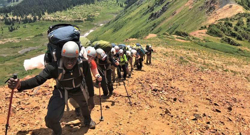 A single-file line of students wearing helmets and carrying backpacks look up at the camera while making their way along a trail.